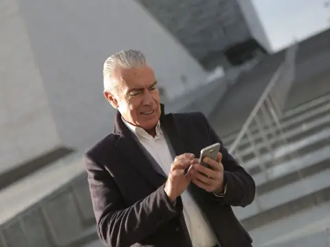 A grey-haired man in a dark coat taps his phone on the steps of a modern building