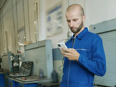 A man in blue work overalls checks his phone on a workshop floor