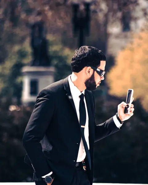 A man in a dark suit and sunglasses walks outdoors, reading his phone