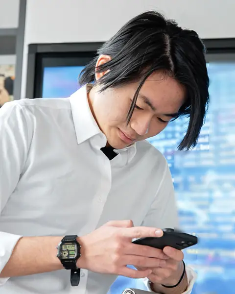 A man in a white shirt checks his phone in an office, in front of a blurred screen of code
